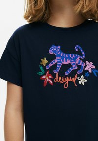 Tricou din bumbac bleumarin, cu un grafic colorat al unui tigru mergător și un design floral în nuanțe de roz, aur și verde, având textul "desigual".