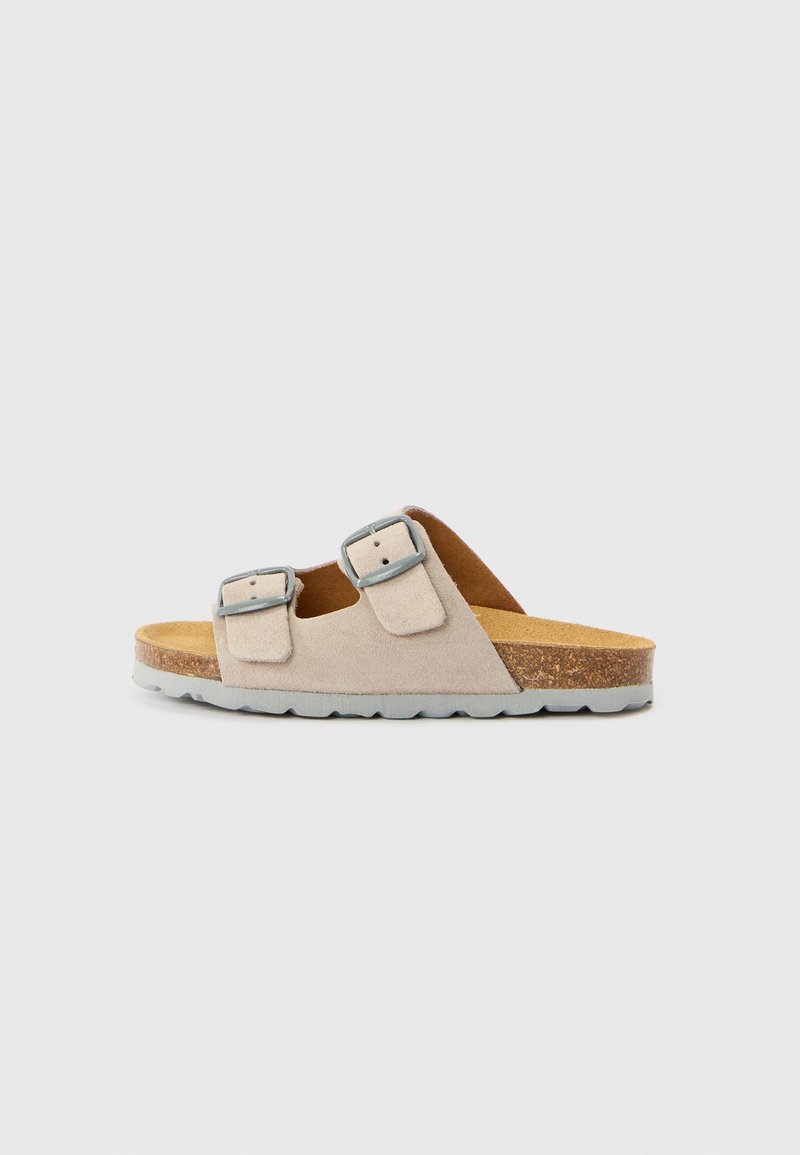 Barna színű velúr szandál két állítható ezüst csattal, parafa talpbetéttel és texturált szürke gumitalppal. Lapos kialakítás, nyitott orr-résszel.