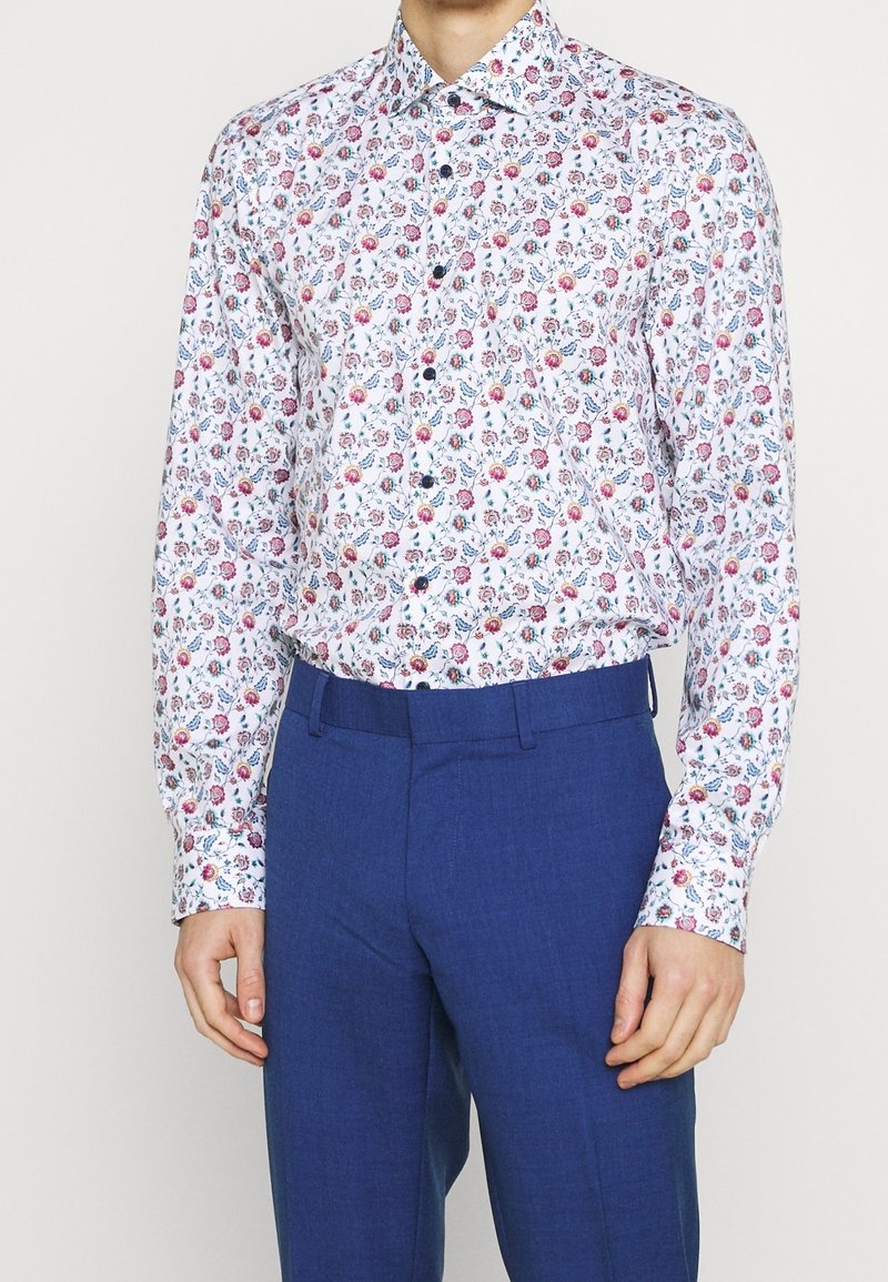 Chemise blanche boutonnée avec un imprimé floral en nuances de rose, bleu et vert. Manches longues, col classique, associée à un pantalon bleu.