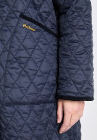 Marineblauwe gewatteerde jas met diamantpatronen, contrasterende zwarte afwerking en een gouden geborduurd logo op de borstzak.
