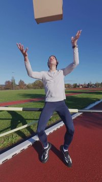 Športnik v svetlo sivi majici z dolgimi rokavi in mornarsko modrih pajkicah, ki na rdečem atletski stezi na prostem žonglira z rjavo kartonsko škatlo.