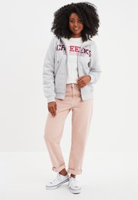 Sudadera gris con cremallera y forro blanco, con texto rojo. Camiseta blanca debajo y pantalones cortos de color rosa pálido. Zapatillas blancas con detalles en negro.