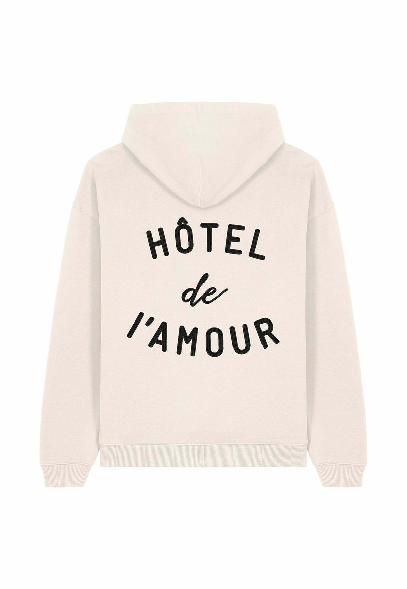 Hoodie color beige chiaro con una vestibilità rilassata, caratterizzato da testo nero che recita "HÔTEL de l'amour" in un design curvo sulla parte posteriore. Texture del tessuto morbida.