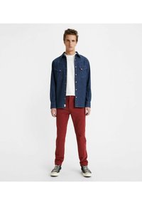 Camicia di jeans blu scuro con due taschini sul petto, abbinata a pantaloni rossi slim-fit. Sneakers in bianco e nero completano il look.