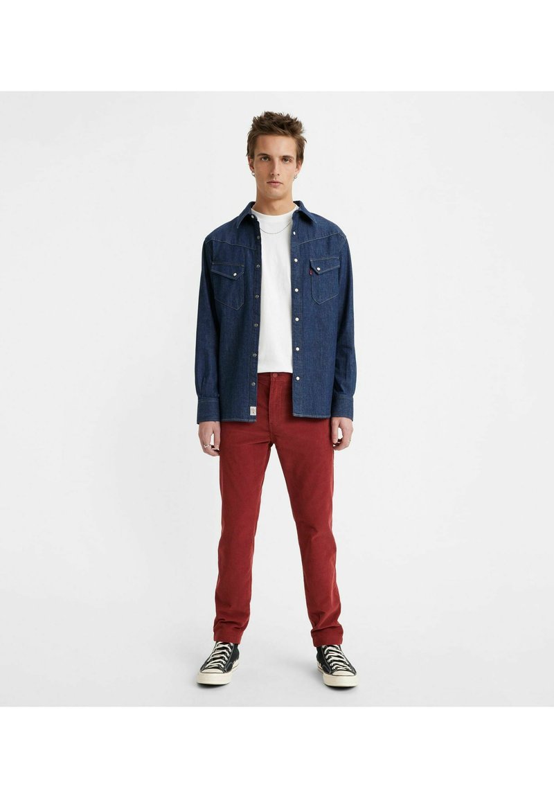 Camicia di jeans blu scuro con due taschini sul petto, abbinata a pantaloni rossi slim-fit. Sneakers in bianco e nero completano il look.