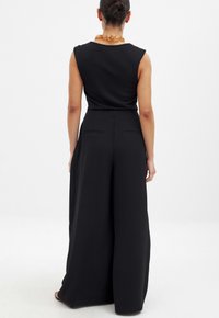 Zwart jumpsuit met mouwloos ontwerp, wijde pijpen en twee achterzakken. Gladde textuur; heeft een ronde halslijn en minimale accenten.