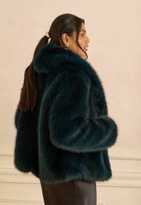 Mākslīgā kažokāda dziļā tumši zilā krāsā ar pūkainu, teksturētu apdari. Īpašības ietver plašu apkakli un brīvu piegriezumu, ar vieglu spīdumu un mīkstu krišanu.