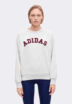 Une femme pose avec un sweat-shirt Adidas gris clair à l’inscription bordeaux et un legging bleu marine, sur un fond clair uni.
