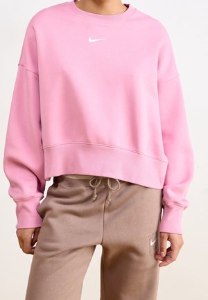 Person trägt ein rosa cropped Nike-Sweatshirt und beige Jogginghose mit Kordelzug und Nike-Logo auf dem linken Oberschenkel.