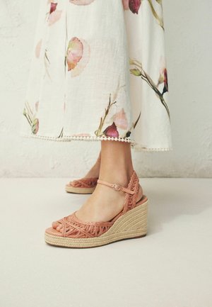 Füße, die rosa gewebte Keilsandalen mit Knöchelriemen tragen, kombiniert mit einem weißen Midikleid mit Blumenmuster und spitzenbesetztem Saum.