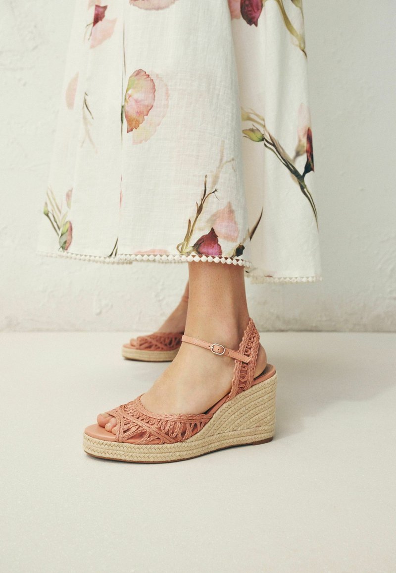 Pieds chaussés de sandales compensées tissées roses avec des brides à la cheville, assorties à une robe midi blanche à fleurs avec un ourlet bordé de dentelle.