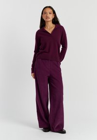 Violetter Pullover mit Kragen und frontaler Knopfdetaillierung, kombiniert mit weiten, violetten Cord-Hosen. Sanfte Textur über das gesamte Kleidungsstück.