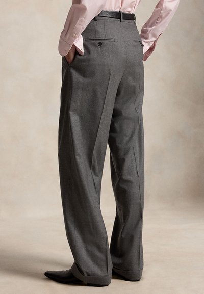 Person trägt graue Hose mit weitem Bein und Gesäßtaschen, schwarzen Gürtel, pinkes langärmeliges Hemd, Hände in den Taschen, steht vor neutralem Hintergrund.