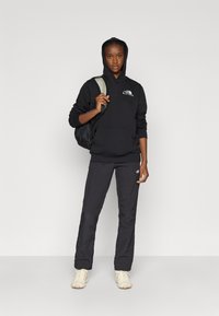 Μαύρο φούτερ με κουκούλα και λογότυπο, μαύρο παντελόνι και μπεζ αθλητικά παπούτσια. Το μοντέλο κρατά ένα μαύρο σακίδιο πλάτης. Απλή και λειτουργική εξωτερική ένδυση.