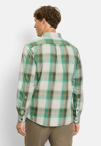 Ein Mann, der von hinten zu sehen ist, trägt ein langärmliges Hemd mit grün-weißen und braunen Karos sowie eine braune Hose vor einem schlichten weißen Hintergrund.