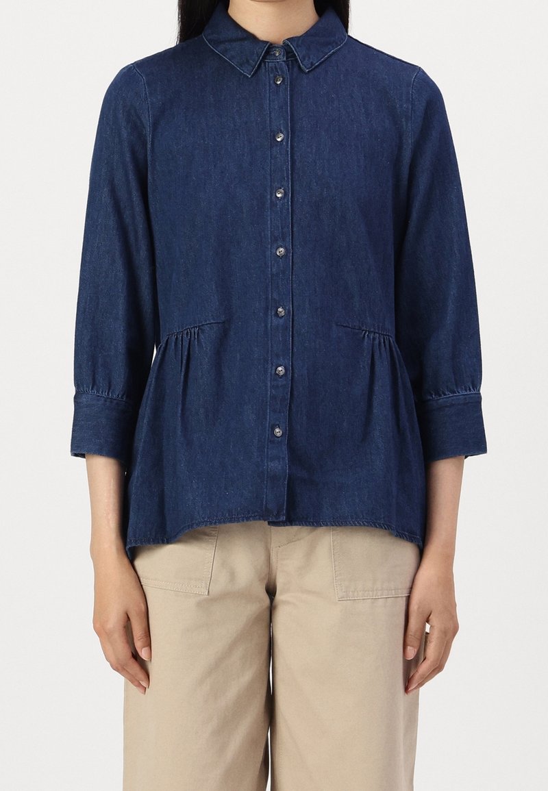 Femme portant un chemisier en denim bleu foncé à boutons avec manches 3/4 et un pantalon beige, debout devant un fond blanc uni.