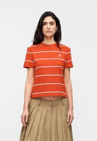 Κόκκινη κοντομάνικη μπλούζα με λευκές ρίγες και μικρό λογότυπο στο στήθος, συνδυασμένη με μπεζ φούστα με πιέτες και ψηλή μέση.