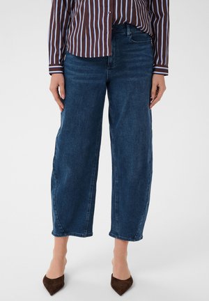 Femme portant un jean large court bleu foncé, une chemise rayée marron et blanche et des chaussures pointues marron foncé, debout sur fond blanc.