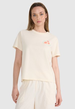 Młoda kobieta w kremowym t-shircie z krótkim rękawem z małym pomarańczowym kwiatowym wzorem i pasujących kremowych spodniach, stojąca na jasnym tle.