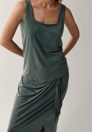 Femme portant un haut sans manches vert foncé et une jupe fronçée assortie avec un nœud sur le côté, debout les mains derrière le dos.