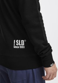 Sweatshirt noir avec des poignets et un ourlet côtelés, arborant un logo brodé blanc "! SLD Since 1993" sur le côté gauche. Texture douce.