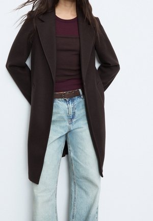 Kobieta ubrana w ciemnobrązowy długi płaszcz na bordowej bluzce i jasnoniebieskich jeansach z wysokim stanem z brązowym paskiem, stojąca na jednolitym tle.