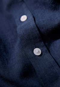 Granatowa lniana koszula wyróżniająca się gładką teksturą, z pionowym zapięciem i białym plastikowym guzikiem. Zbliżenie na materiał i guzik.