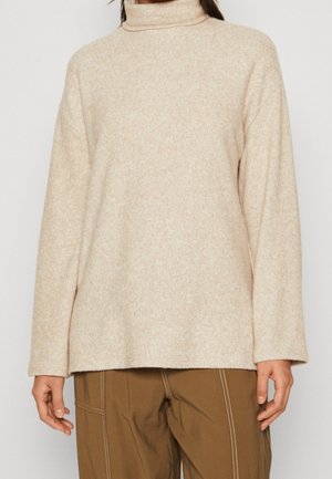 Personne portant un pull à col roulé beige à manches longues, associé à un pantalon marron avec des coutures visibles et des poches.