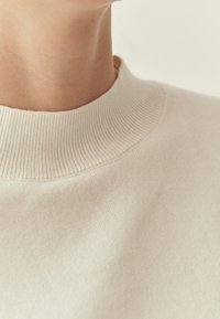 Pull couleur crème avec un col côtelé et une texture lisse, mise au point rapprochée sur la zone du cou et les détails du tissu.