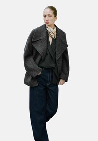 Abrigo de lana gris con solapas sobredimensionadas, combinado con una blusa negra y pantalones de mezclilla oscura holgados; presenta una bufanda ligera de patrón alrededor del cuello.