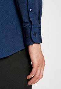 Mörkblå skjorta med strukturerad textil, med ett subtilt prickmönster, knappärmar och skräddarsydd passform. Hand vilande på mörka byxor.