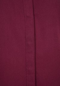 Tessuto bordeaux, superficie liscia con una leggera lucentezza. Cucitura centrale che corre verticalmente, creando un aspetto strutturato. Materiale leggero.