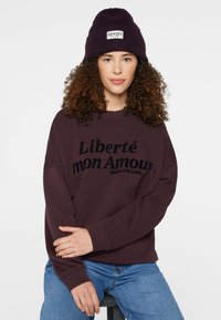Maroon sweatshirt with "Liberté mon Amour" printed in black, paired with a dark purple knit beanie featuring a sewn label. Blue jeans visible.