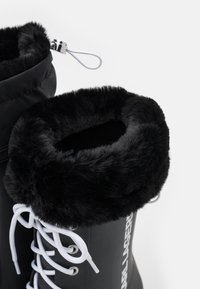 Black waterproof boots with a faux fur cuff, white laces, and a smooth finish. Features a drawstring closure and embossed logo detail.