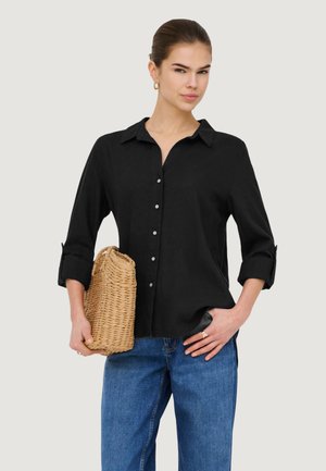 Donna che indossa una camicia nera abbottonata e jeans blu, tenendo in una mano una pochette intrecciata in paglia.