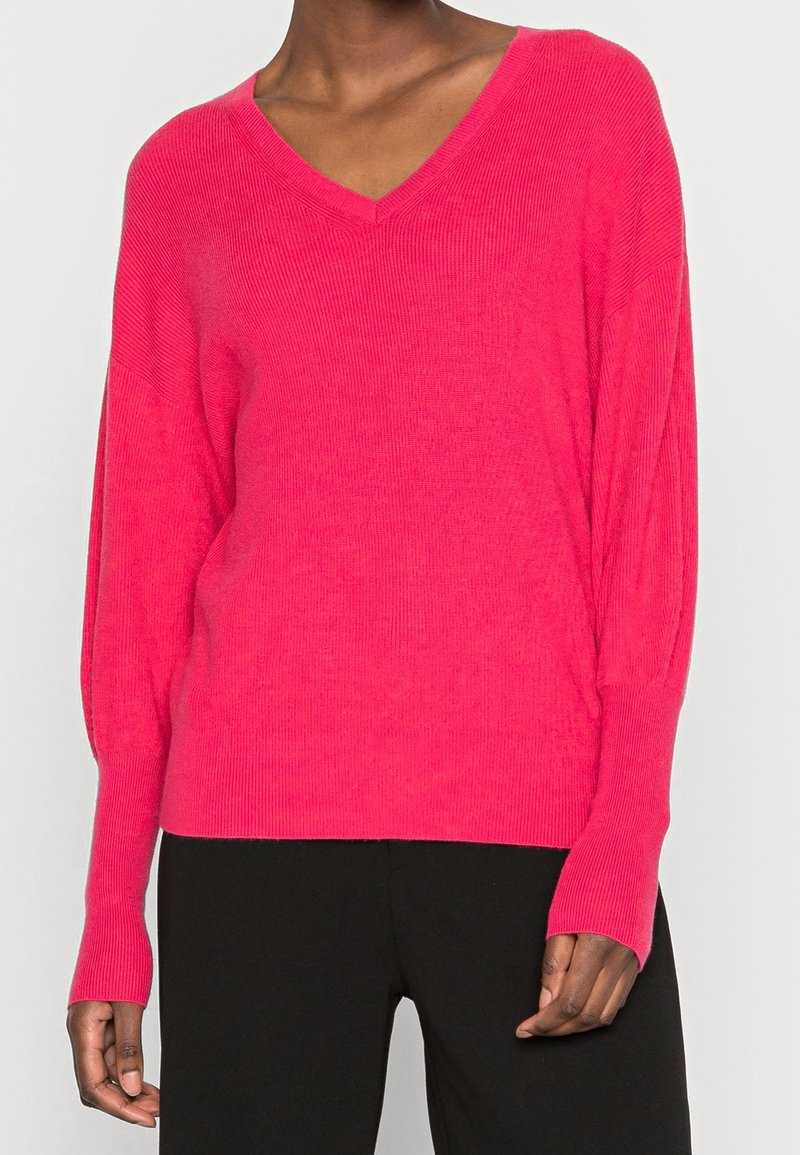 Personne portant un pull à manches longues rose vif col en V et un pantalon noir, debout devant un fond clair uni.