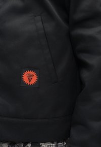Μαύρο μπουφάν από ματ ύφασμα με κόκκινο και μαύρο έμβλημα λογότυπου που παρουσιάζει σχεδιασμό ακτίνων ήλιου. Περιλαμβάνει πλάγια τσέπη με γωνία.