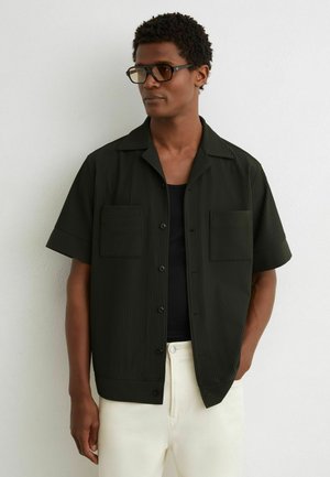 Mann mit kurzen lockigen Haaren, der ein schwarzes, geripptes Kurzarmshirt, ein schwarzes Tanktop, weiße Hose und rechteckige Brille trägt, nach rechts blickend.