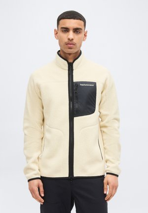 Homme portant une veste polaire crème avec une fermeture éclair noire et une poche poitrine étiquetée "Peak Performance", debout devant un fond blanc uni.
