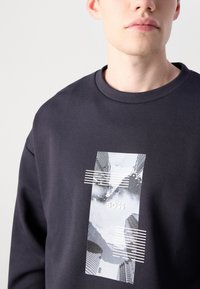 Felpa blu scuro con scollatura rotonda presenta una grafica che mostra paesaggi astratti e il testo "BOSS" in bianco, accentuato da linee a strisce.
