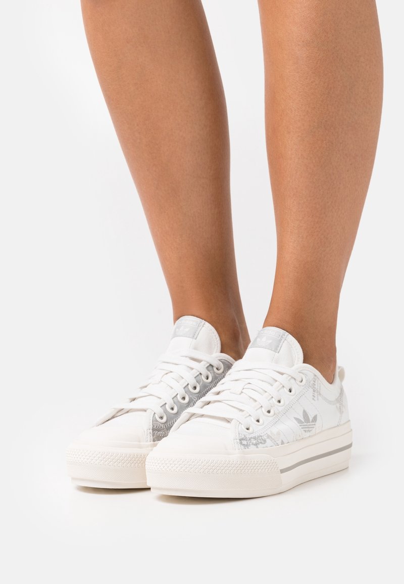 White low-top Adidas sneakers with gray logo patterns worn on bare legs against a white background.