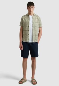Chemise à manches courtes de couleur vert clair avec un motif de vagues, associée à un short bleu marine. Le modèle porte des sandales grises. Design simple et décontracté.