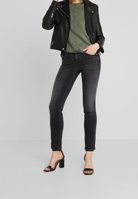 Mujer con chaqueta de cuero negro, camiseta verde, vaqueros ajustados negros y sandalias de tacón abiertas negras de pie con una mano en el bolsillo.