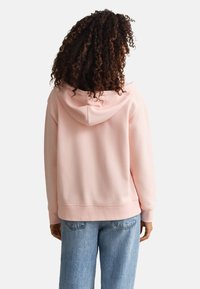 Osoba z kręconymi włosami ubrana w jasnoróżową bluzę z kapturem i niebieskie dżinsy, zwrócona tyłem na jednolitym białym tle.