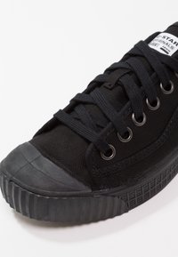Baskets basses noires avec embout en caoutchouc et design à lacets, avec œillets métalliques et une étiquette sur la languette avec le texte de la marque.