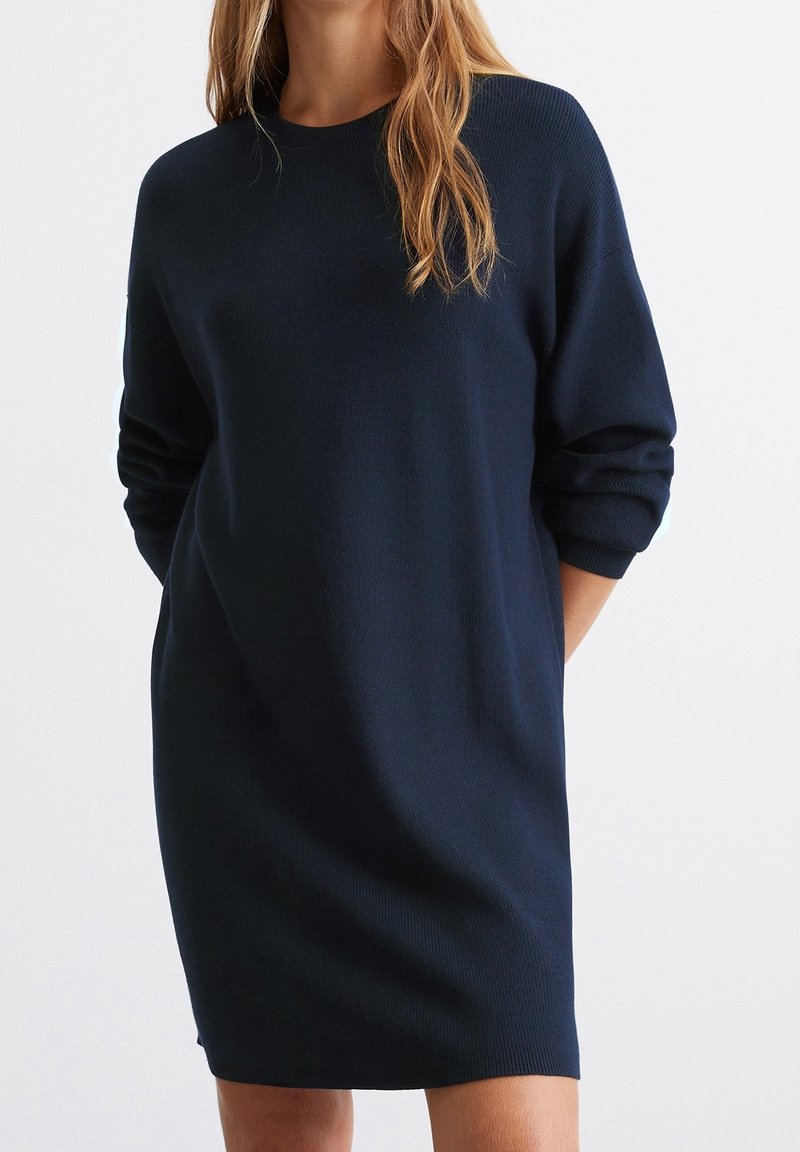 Femme portant une robe-pull en maille bleu marine à manches longues, debout, les mains dans le dos, devant un fond blanc uni.