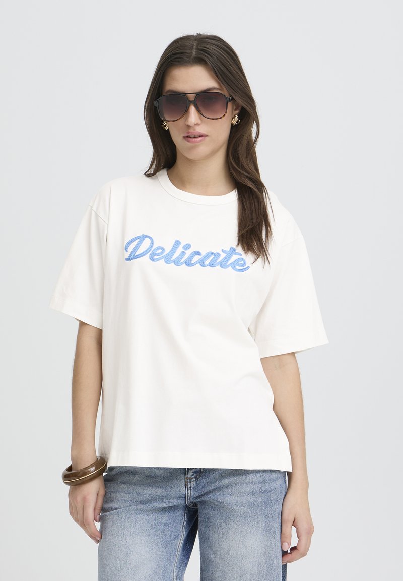 T-shirt crème à manches courtes avec "Delicate" brodé en script bleu clair. Portée avec un jean bleu délavé et un bracelet brun épais.