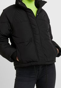 Černá péřová bunda se zipem vpředu, vysokým límcem a dvěma předními kapsami. Má šité panely a mírně oversize střih.