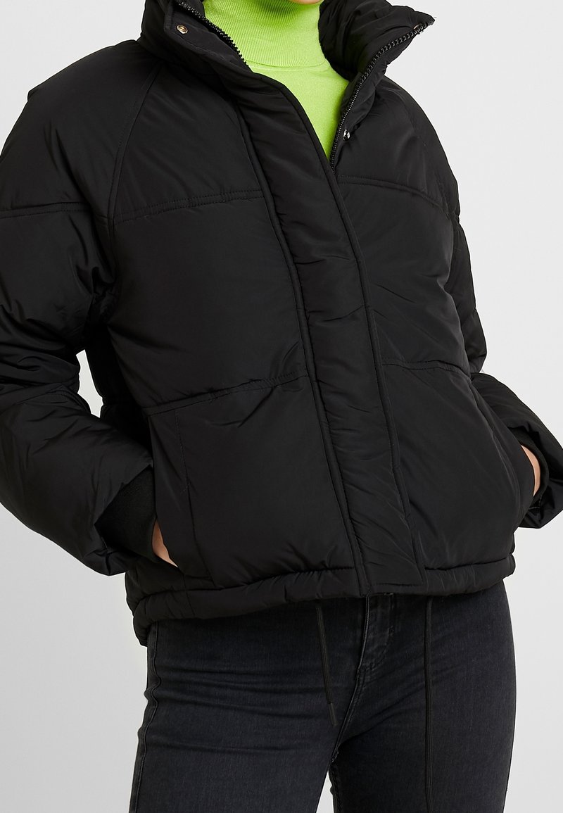Černá péřová bunda se zipem vpředu, vysokým límcem a dvěma předními kapsami. Má šité panely a mírně oversize střih.