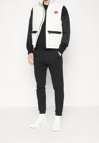 Weiß gesteppte Weste mit schwarzen Akzenten, Reißverschlusstaschen und einem roten Logo, getragen über einem schwarzen Pullover und schwarzen, schmal zulaufenden Jogginghosen, mit weißen Sneakern.